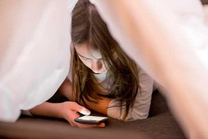 young girl hiding under covers looking at her phone