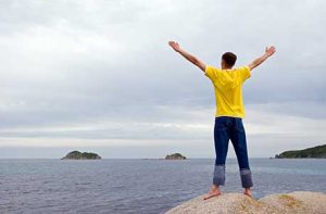 Raising Teenagers - White male teenager with arms open wide and back to camera gazes at the ocean